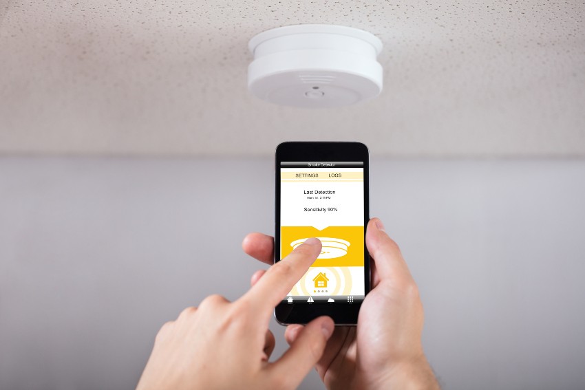 Mittels Handy konfiguriert eine Person einen Smart-Home-Rauchwarnmelder, der an der Decke eines Wohnraums angebracht ist